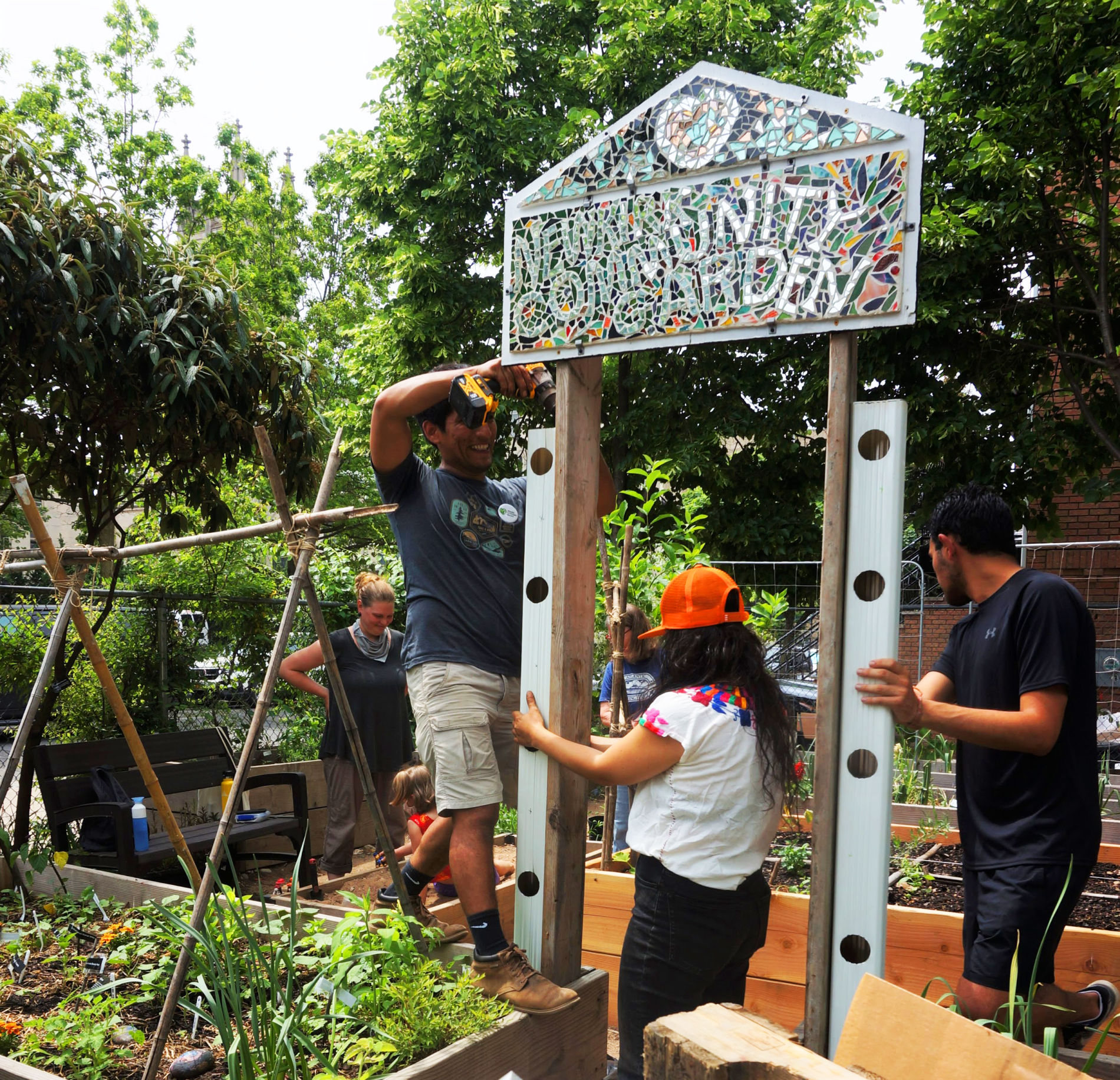 Three volunteers attaching metal vertical planters to the side of a garden arch. On top of the arch is a mosaic sign reading "Newkirk Community Garden."