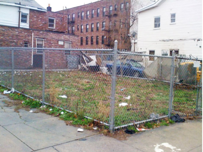 A fenced grassy lot on a residential street, with lots of litter on it