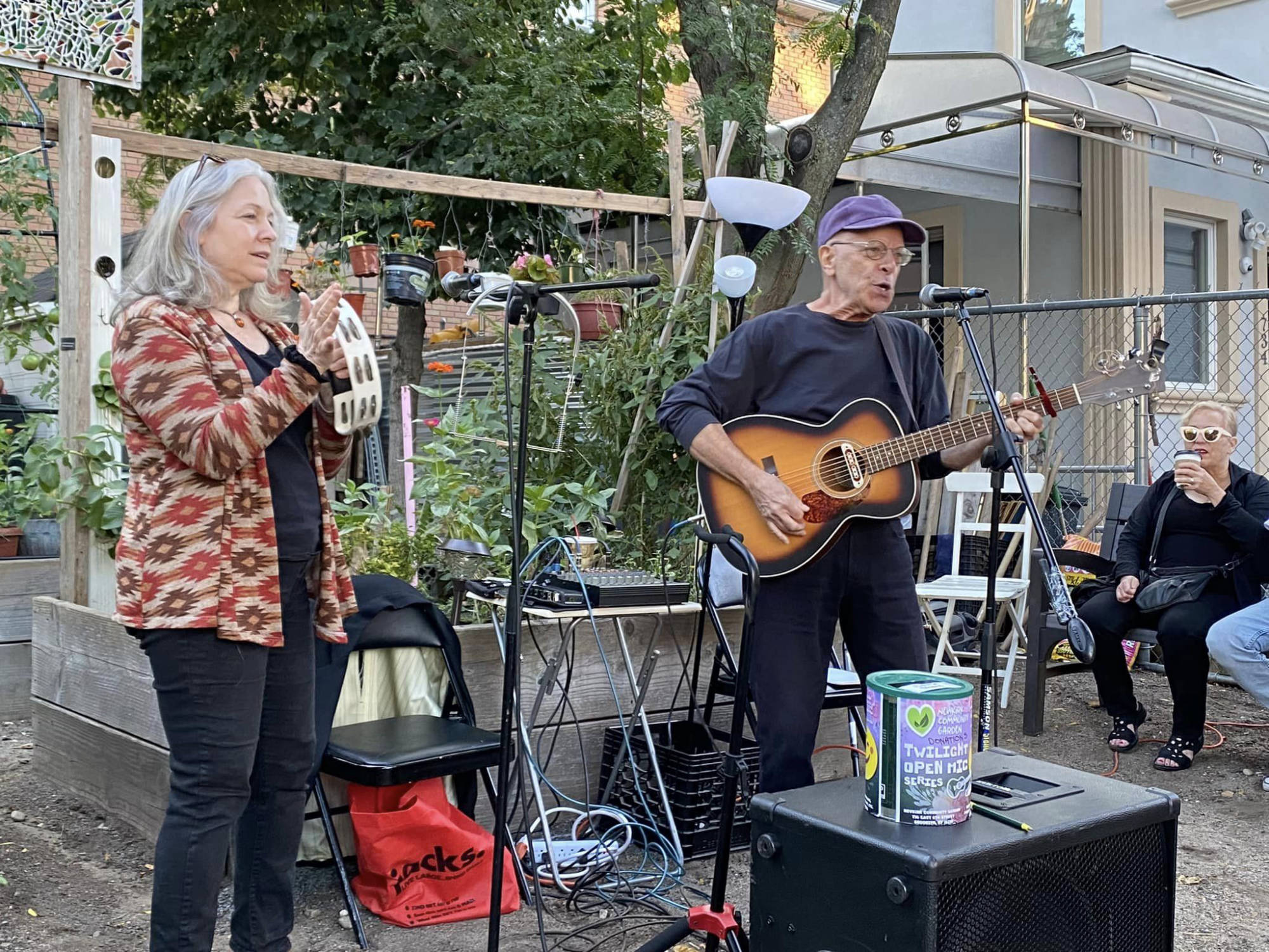 Two people performing a song at open mic night in the garden, with audience members on benches next to them.